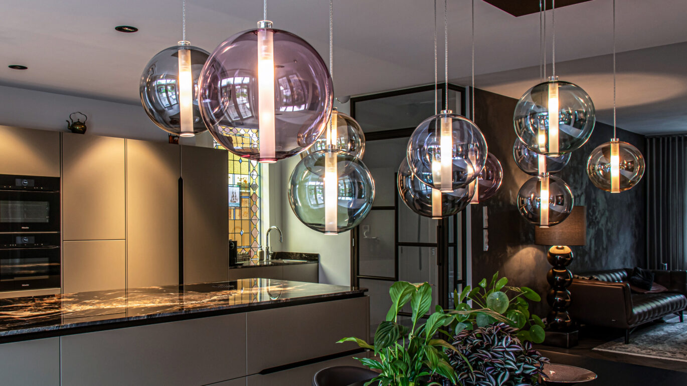 Moderne keuken met hanglampen van Straluma, voorzien van gekleurd glas, boven een donker aanrechtblad, gecombineerd met stijlvolle inrichting en groene planten.