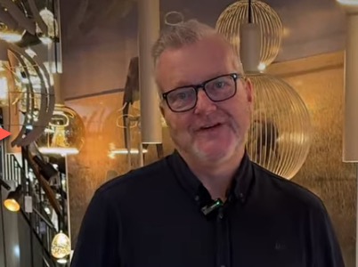 John in de showroom van Straluma, omringd door moderne armaturen in zand-, platinum- en aardetinten; still uit de video over trendkleuren verlichting 2026.